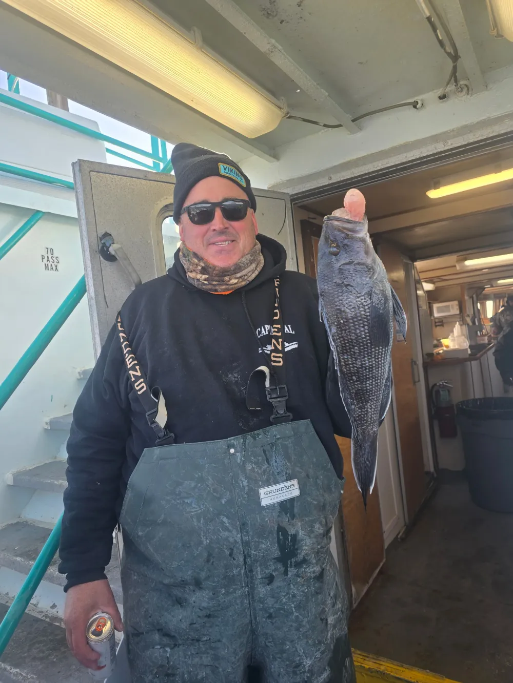 Person in winter clothing holds a fish on a boat, with a can in hand.