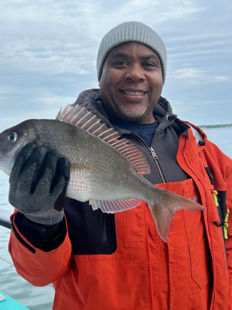 Person in orange jacket holding a fish and smiling on a cloudy day.