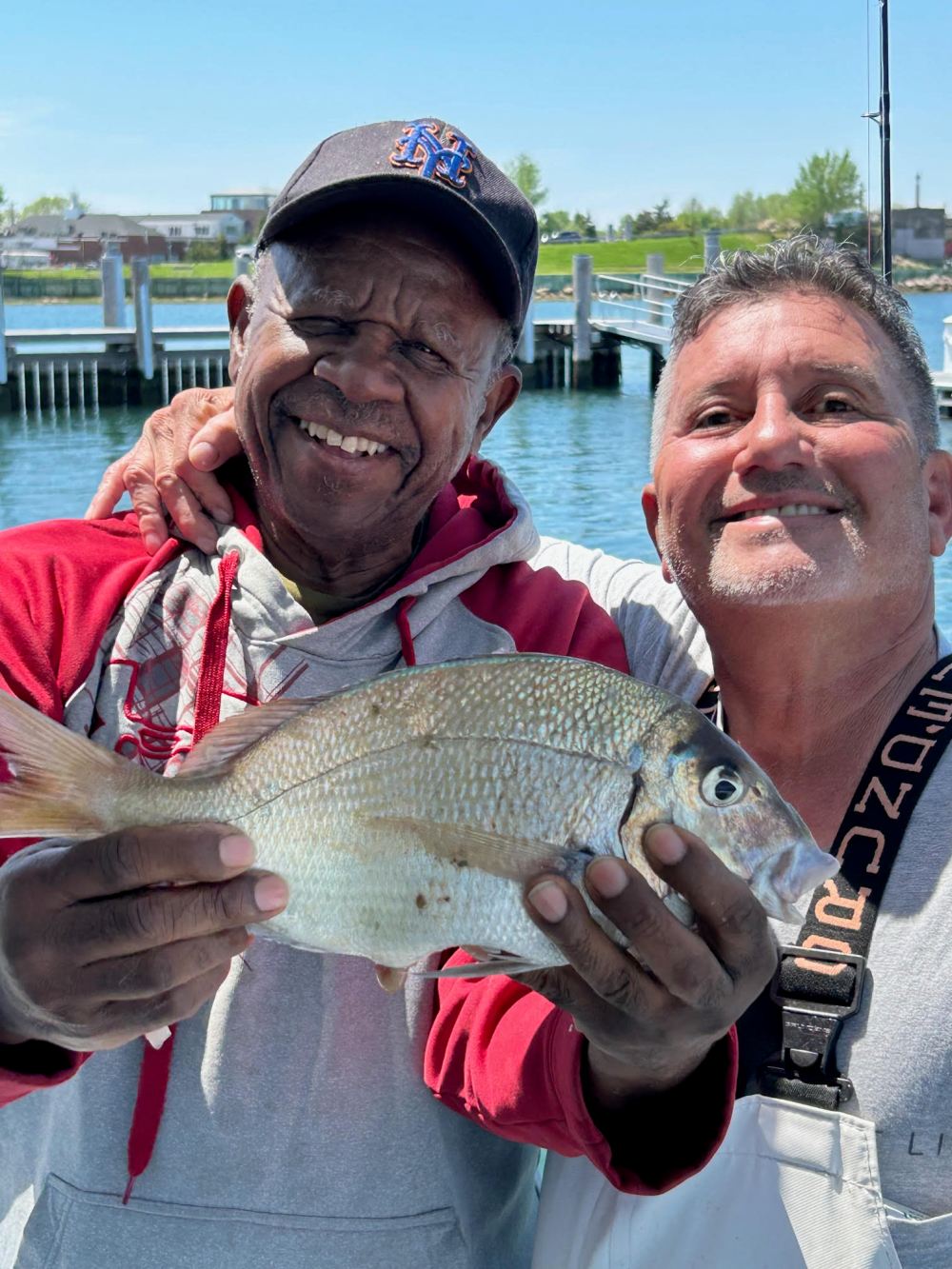 Two men smiling, holding a fish on a dock near water.