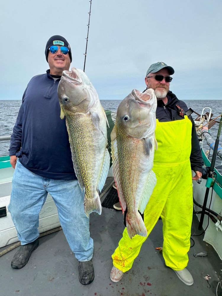 Two men on a boat holding large fish, one in yellow overalls.