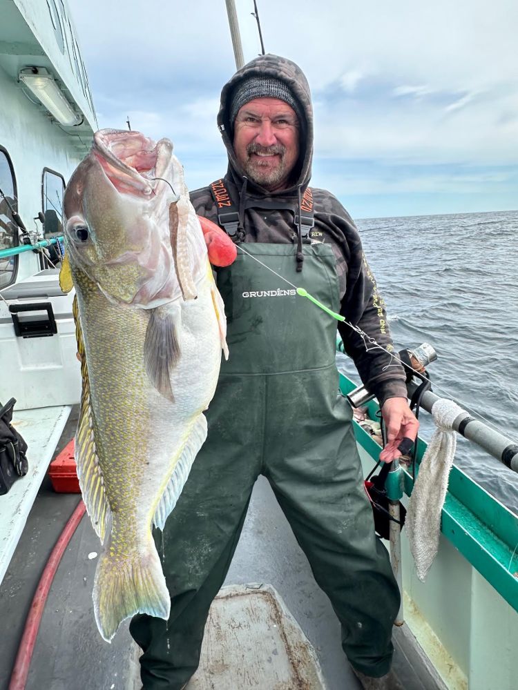 Man on boat holding large fish, wearing green overalls.