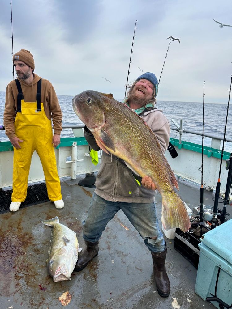 Man on boat holding large fish, another man in yellow overalls watches.