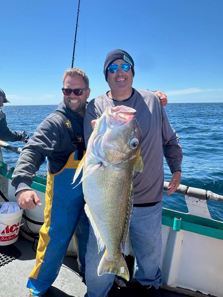 Two men on a boat holding a large fish, smiling under a sunny sky.