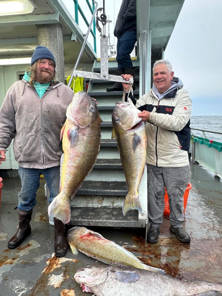 Two people holding large fish on a boat deck with additional fish on the floor.