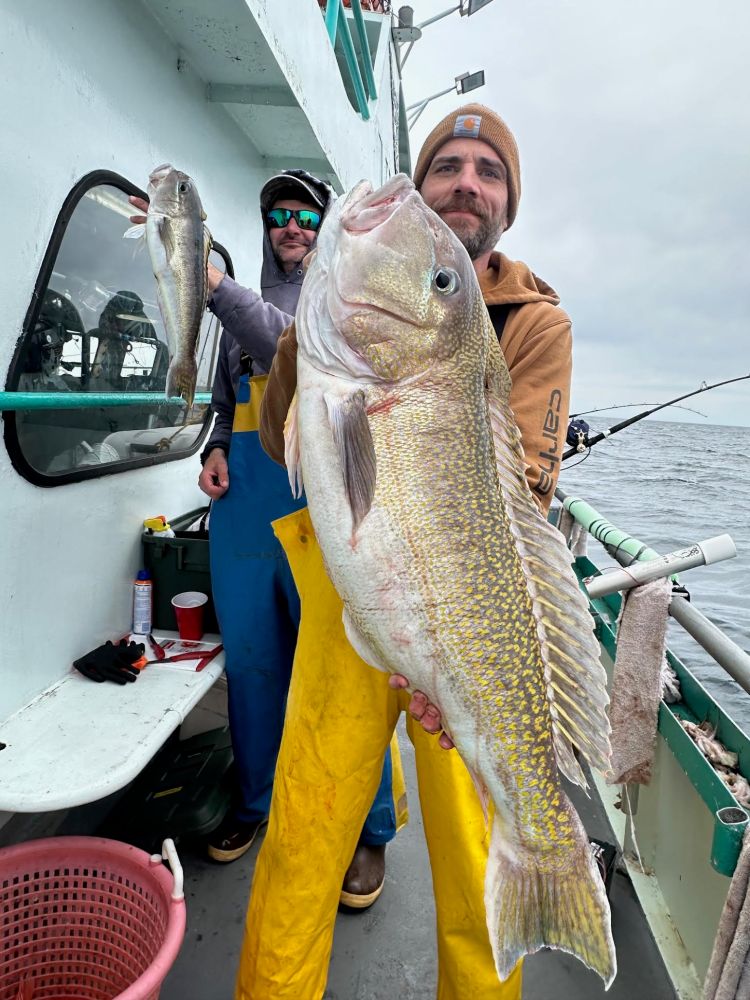 Two people on a boat holding large fish, with one person in focus wearing yellow overalls.