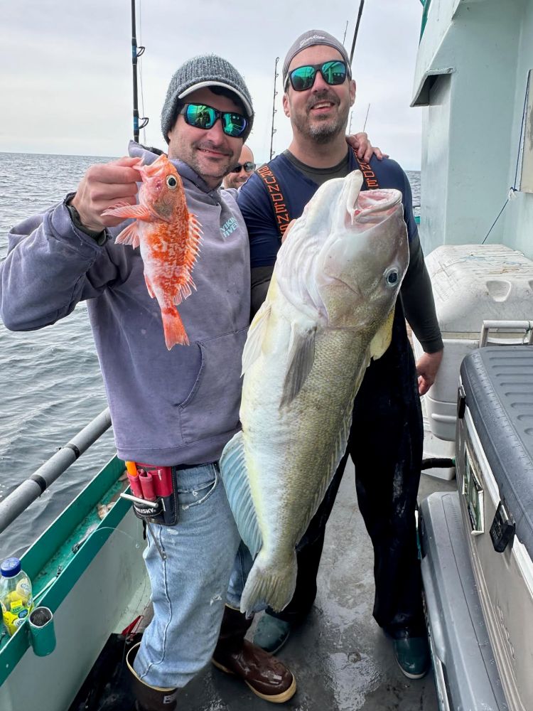Two men on a boat holding large fish, one red and one gray, wearing casual and fishing gear.