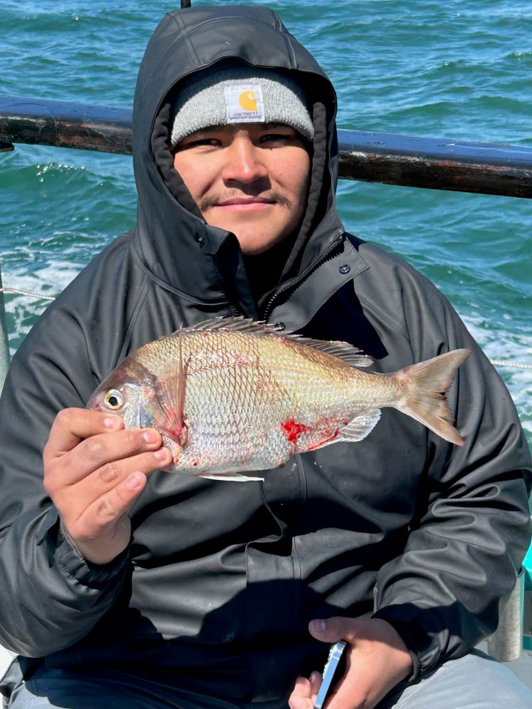 Person in a hooded jacket holding a fish on a boat with ocean in the background.