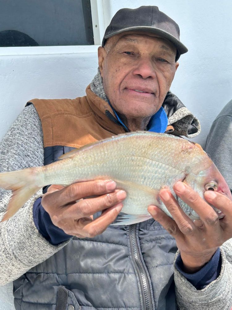 Elderly man holding a large fish, wearing a cap and layered clothing, sitting against a white wall.