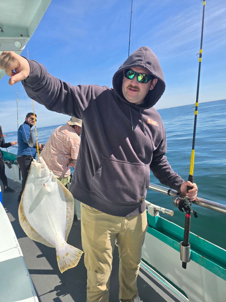 Person in hoodie holding a fishing rod and a large fish on a boat.
