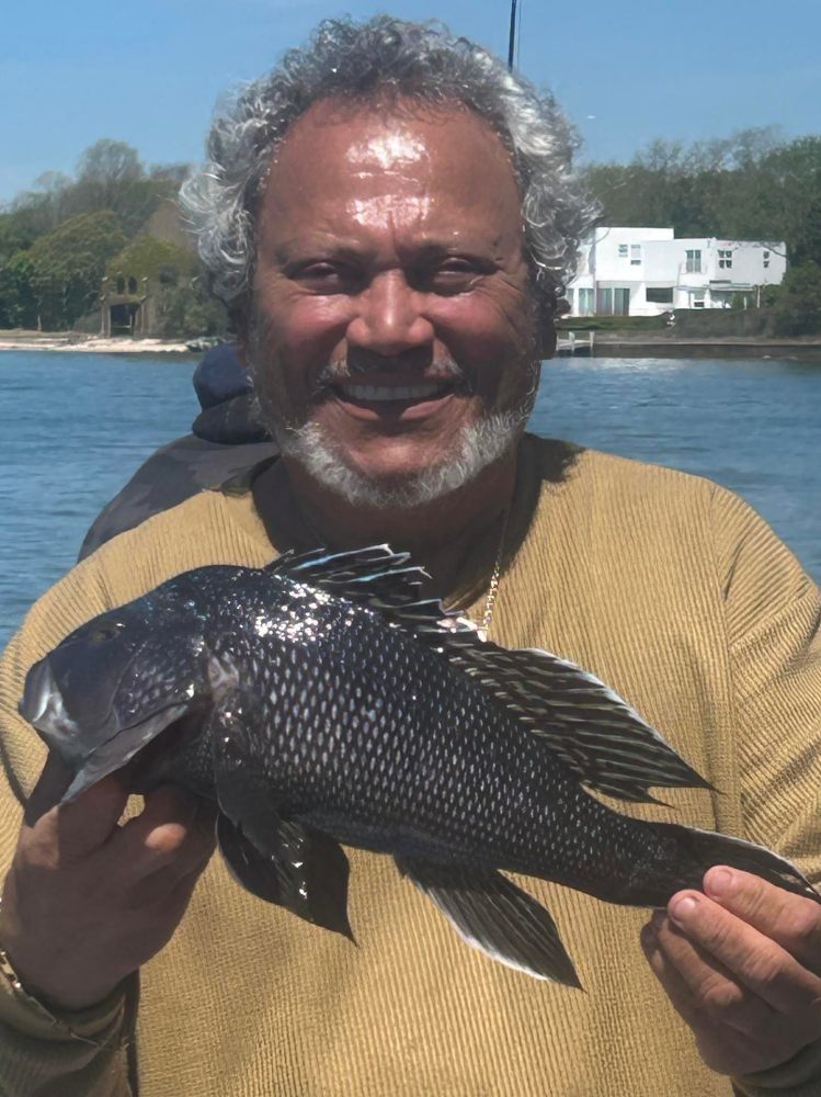 Man holding a fish by the water with houses in the background.