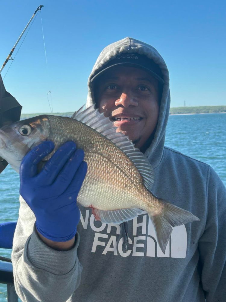 Person in hoodie and gloves holding a fish near a lake.