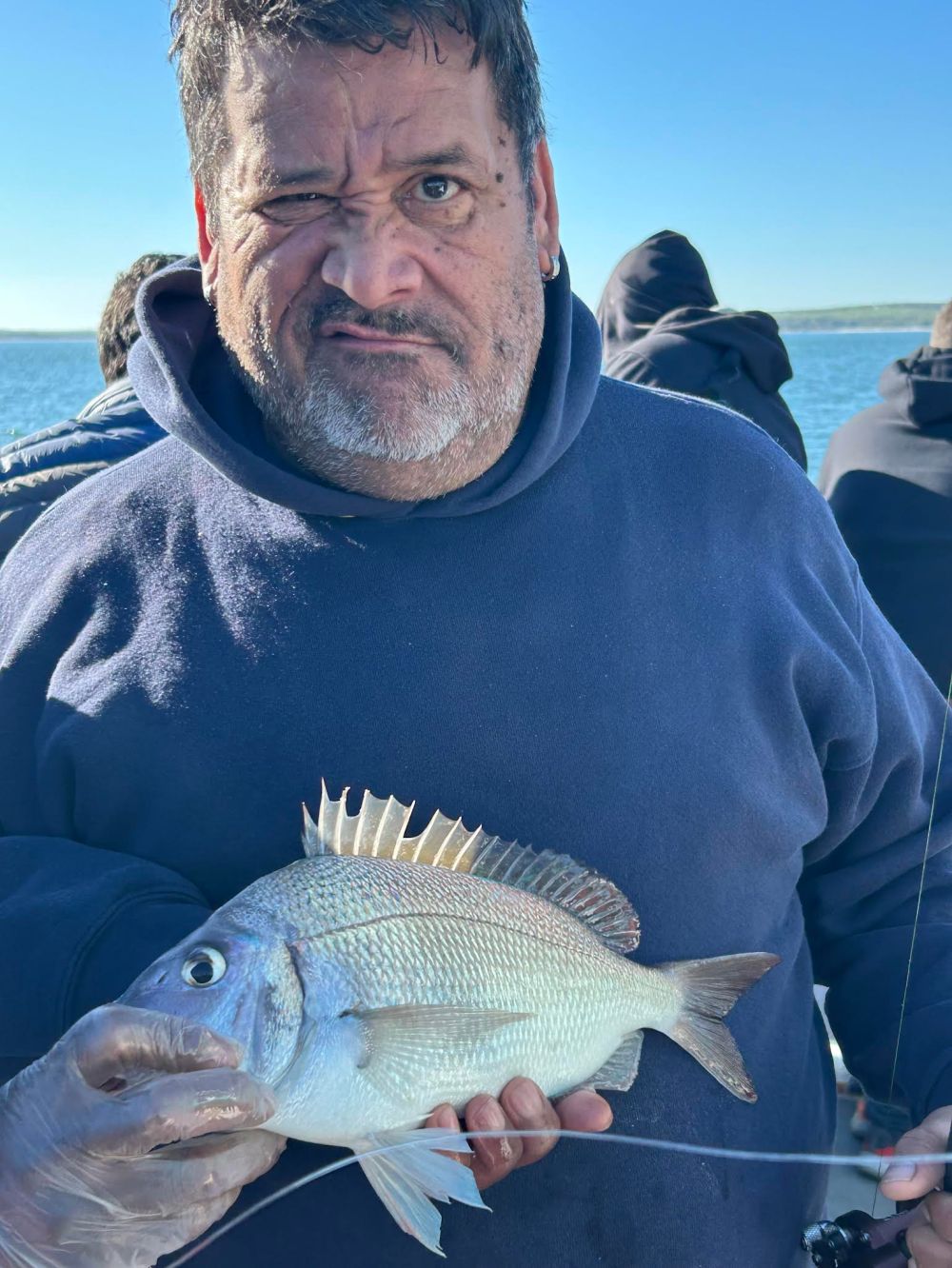 Man in hoodie holding a large fish, squinting at the camera. Clear blue sky and water in background.