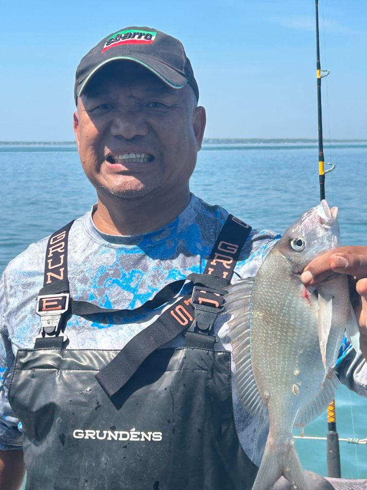 Person holding a fish on a boat, wearing a cap and overalls, with water in the background.