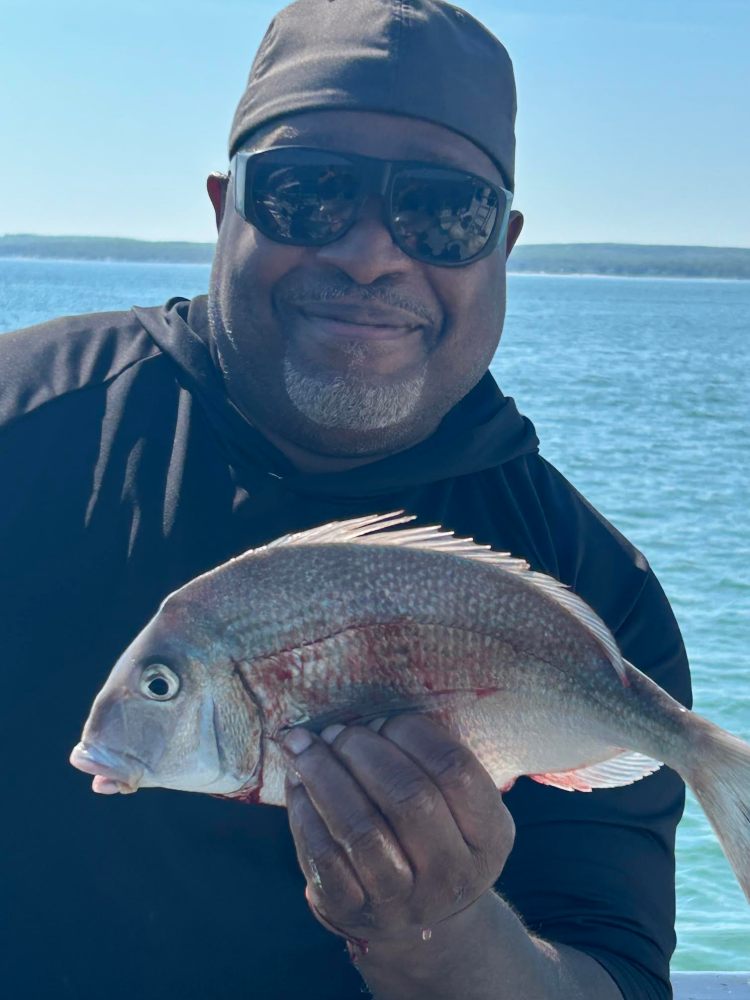 Man in sunglasses holding a freshly caught fish by the water.