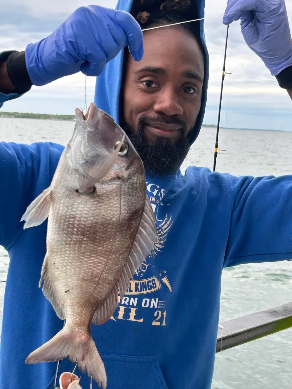 Person in blue hoodie holding a fish on a fishing line by the water.