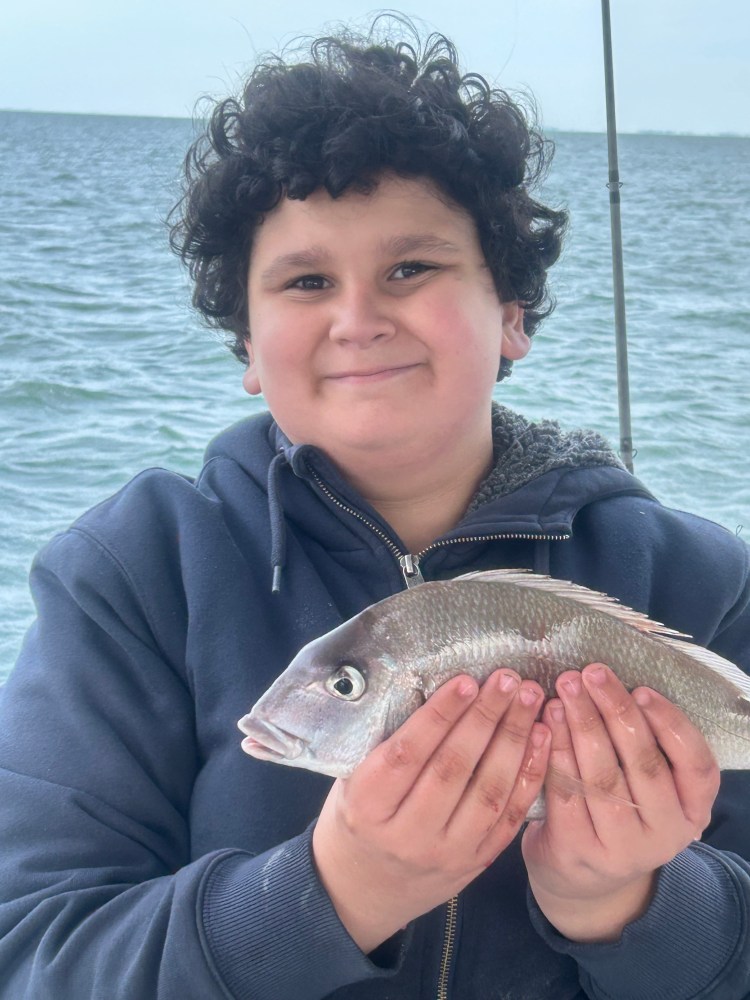 Child smiling and holding a fish on a boat with the sea in the background.