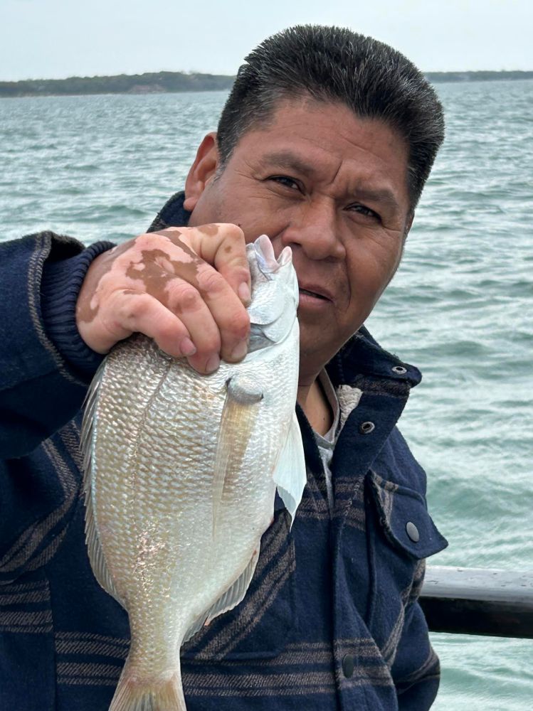 Man holding a fish by the water, wearing a dark jacket.