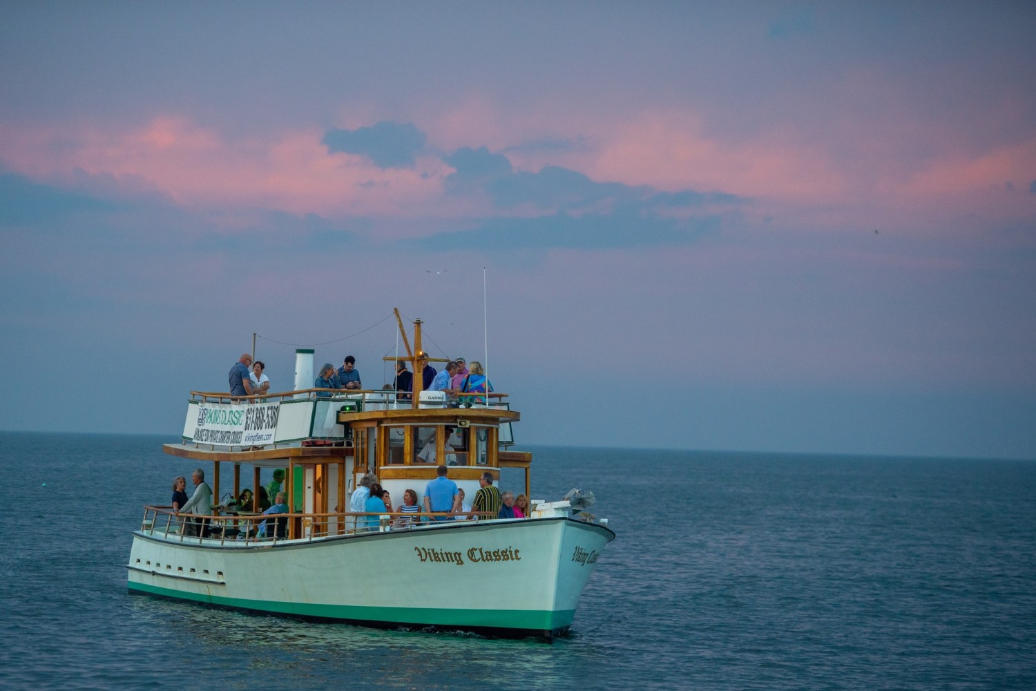 Boat with people on calm sea under a pink sky at dusk.