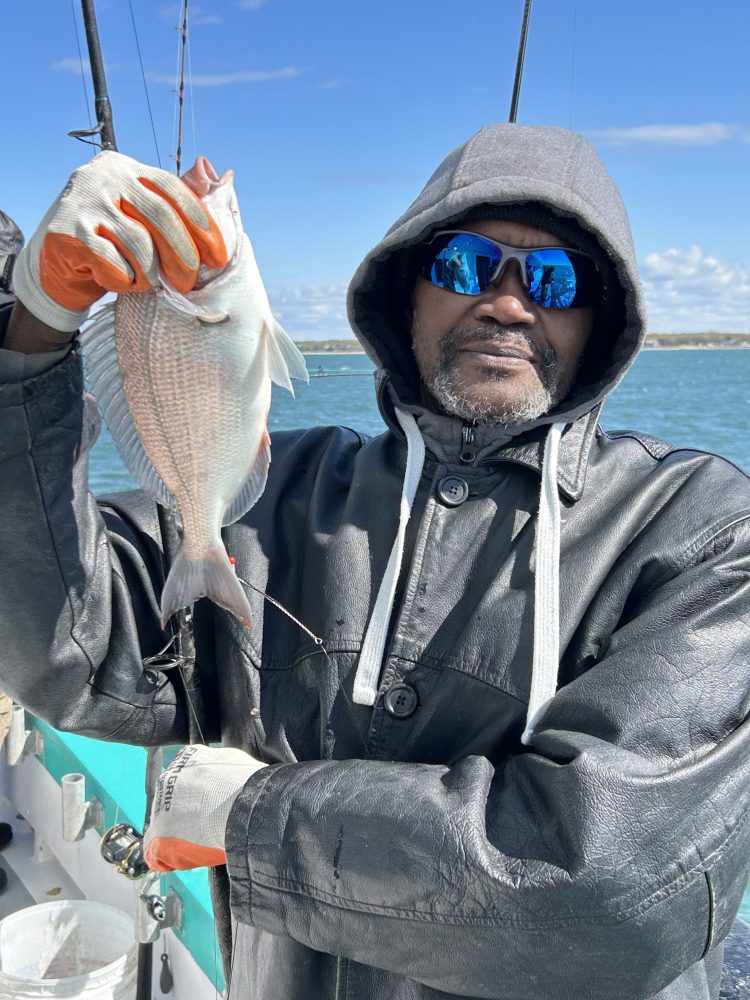 Person in black jacket and sunglasses holding a fish on a boat.