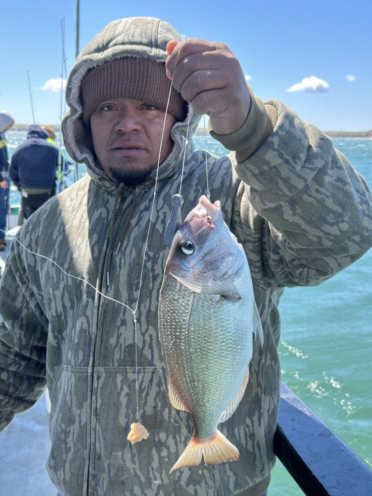 Person in camo hoodie holding a fish on a line by the water.