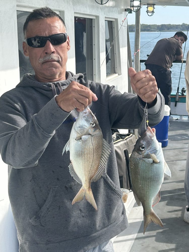 Person holding two fish on a boat, wearing sunglasses and a hoodie.
