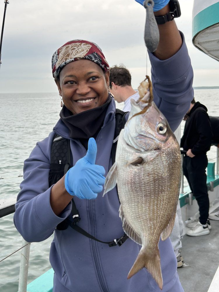 Smiling person holding a fish on a boat, wearing gloves and a headscarf, with people and water in the background.