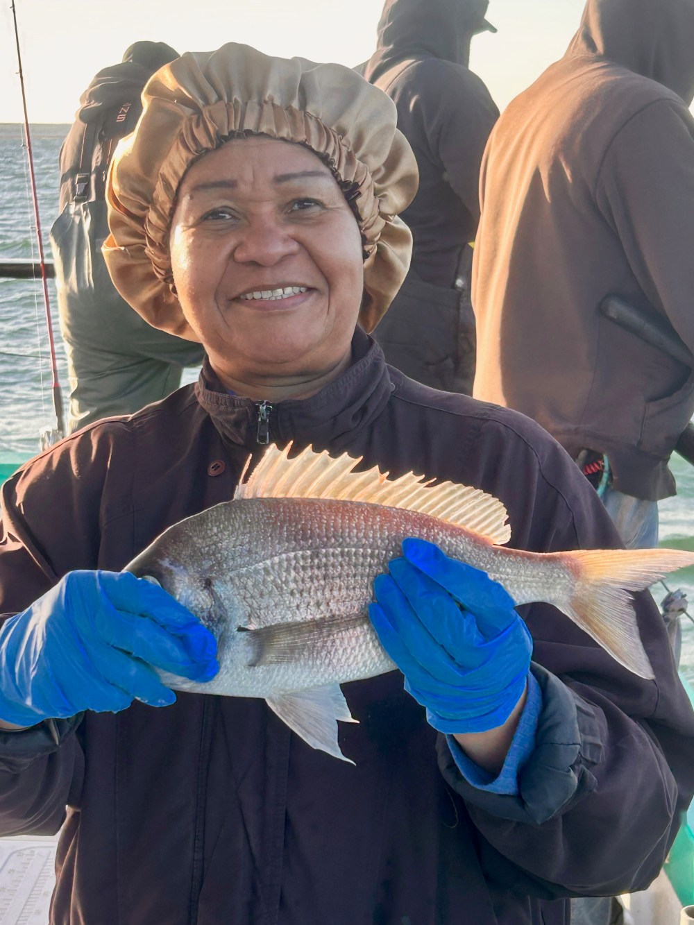 Person smiling, holding a fish on a boat, wearing gloves and a bonnet.