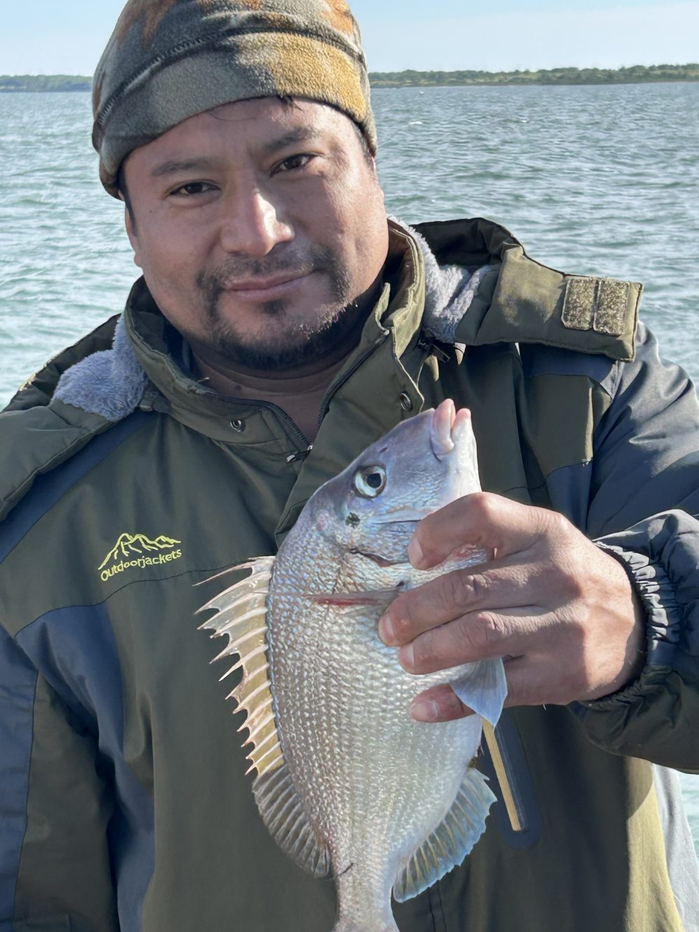 Person holding a fish near a body of water, wearing a jacket and beanie.