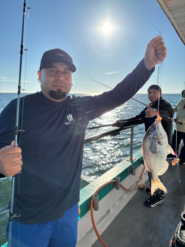 Man holding a fish on a boat with another person in the background, ocean and sunlight visible.