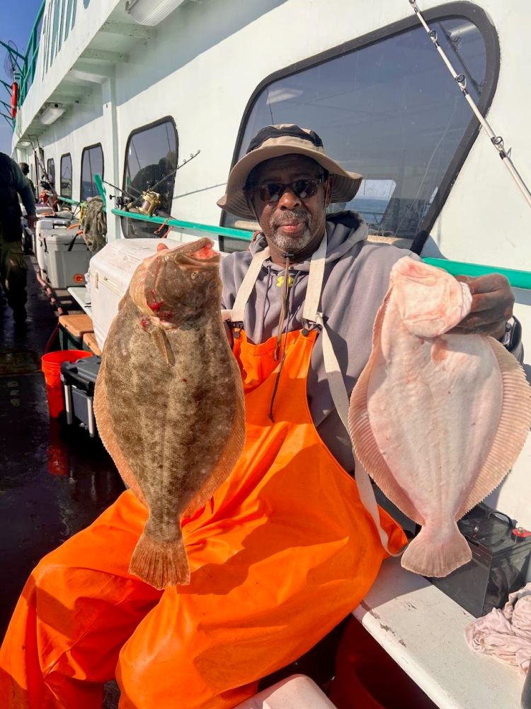 Man on boat holding two large flatfish, wearing orange overalls and hat.