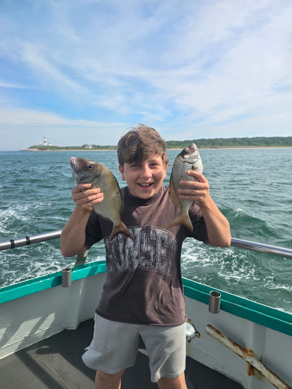 Boy on boat holding two fish, ocean and lighthouse in background.
