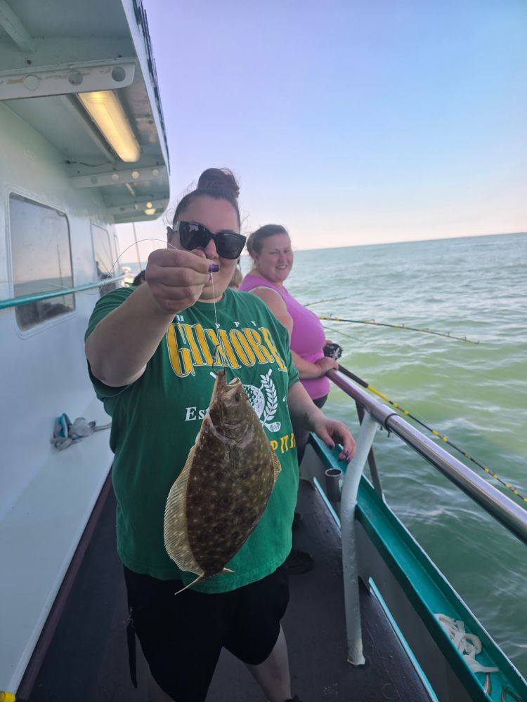 Two people on a boat, one holding a flatfish, with the ocean in the background.