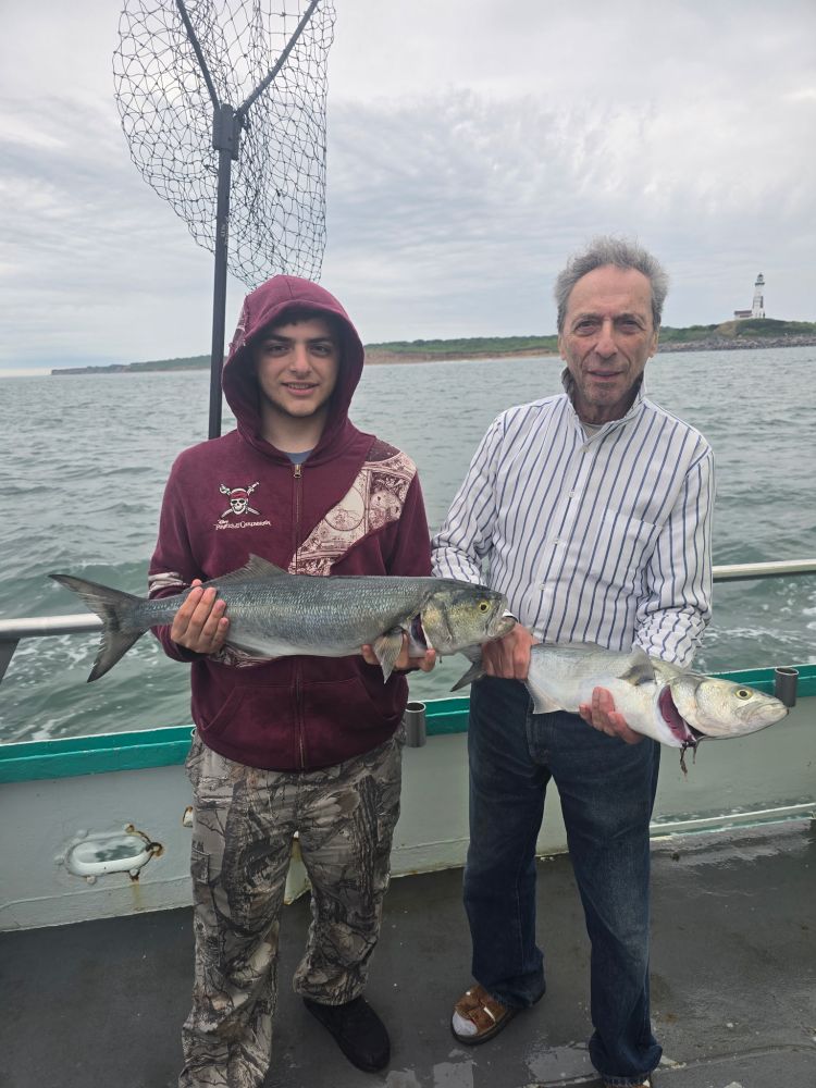 Two people on a boat holding caught fish with a net and lighthouse in the background.