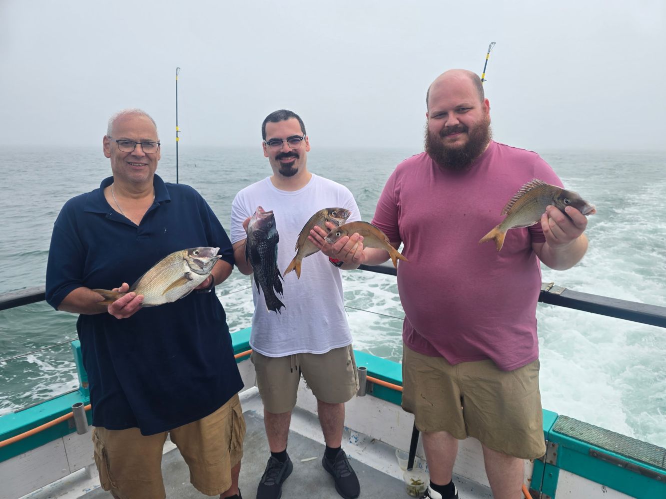 Three men on a boat holding fish with the ocean in the background.