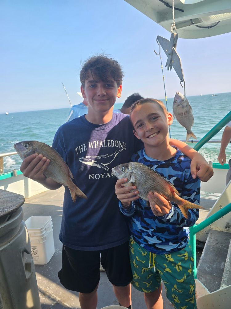 Two boys smiling, holding fish on a boat, with ocean in the background.