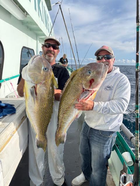 Two people on a boat holding large fish, smiling at the camera.