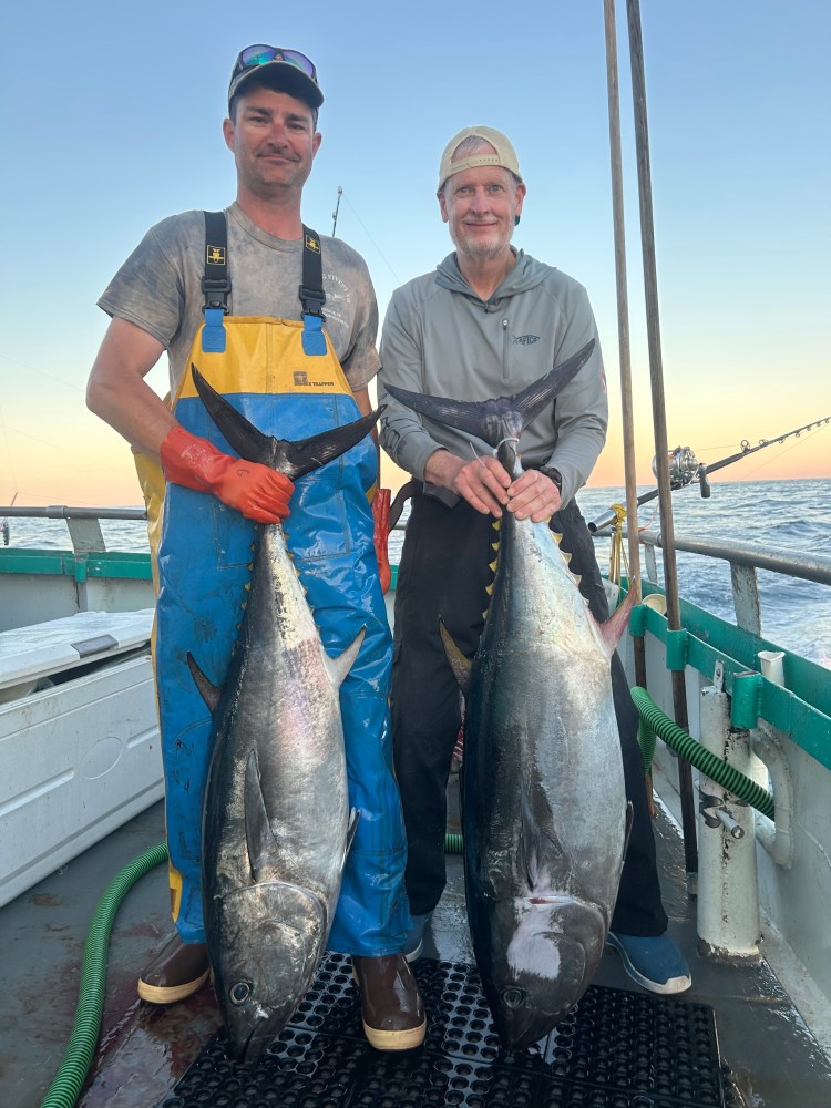 Two men on a boat holding large fish with ocean in background.