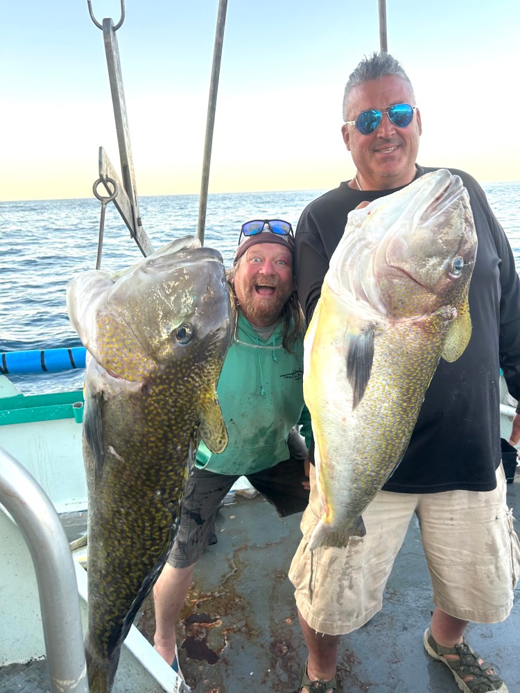 Two men on a boat holding large fish, smiling at the camera.