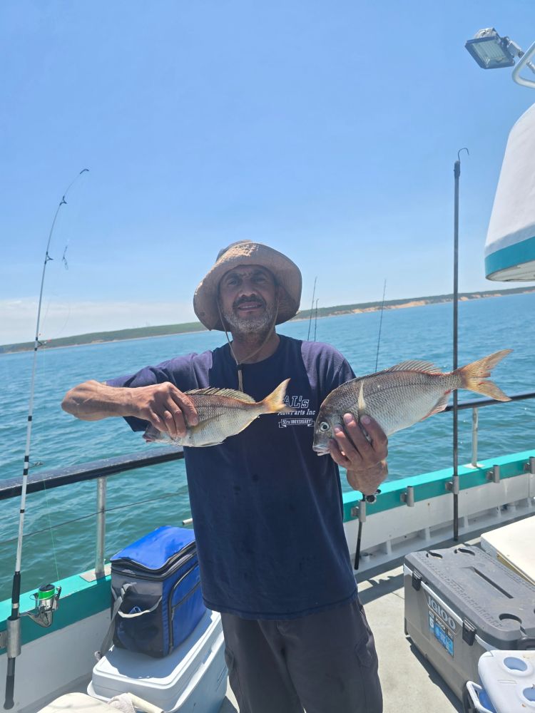 Man on boat holding two fish, with fishing rods and ocean in background.