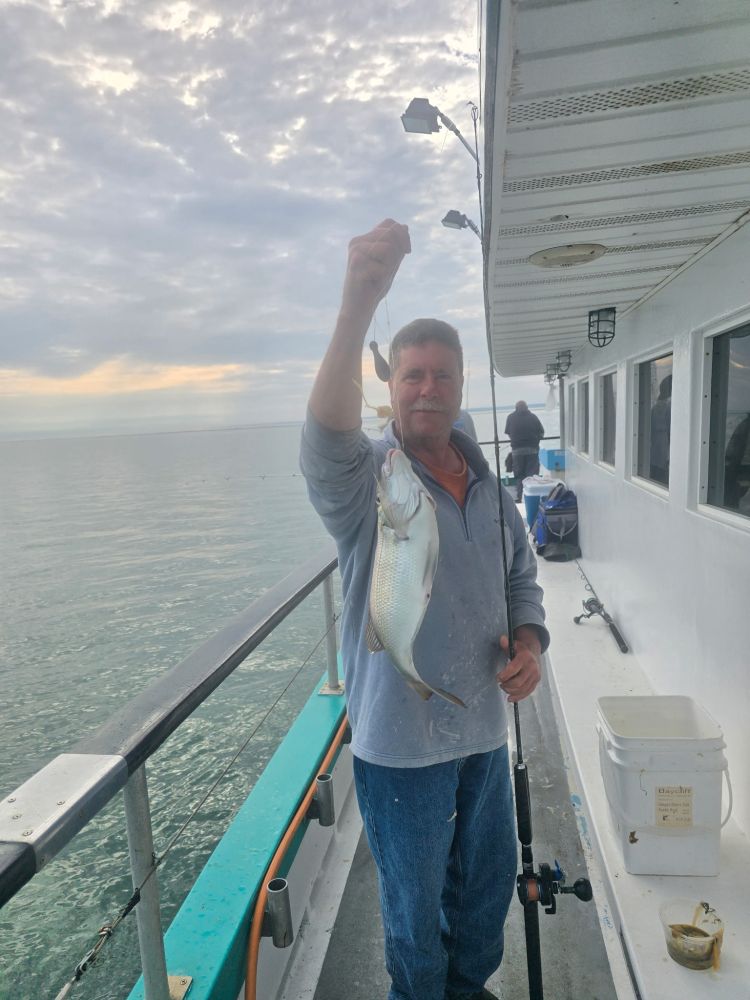Man holds fish on boat deck, with cloudy sky and calm sea in background.