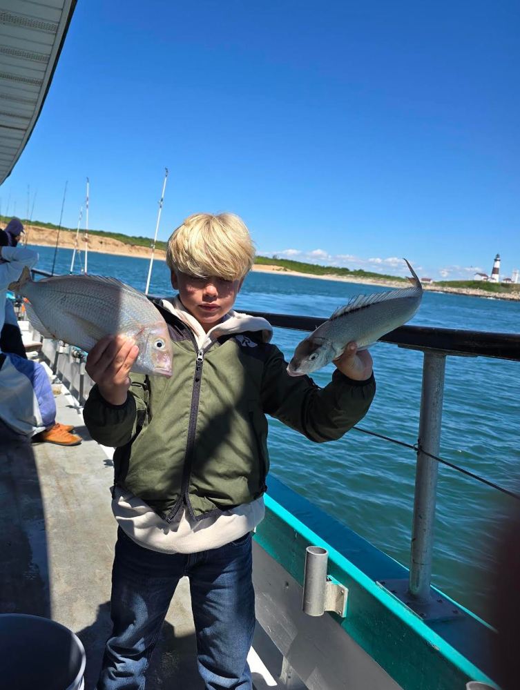Child on boat holding two fish, with blue water and coastline in background.