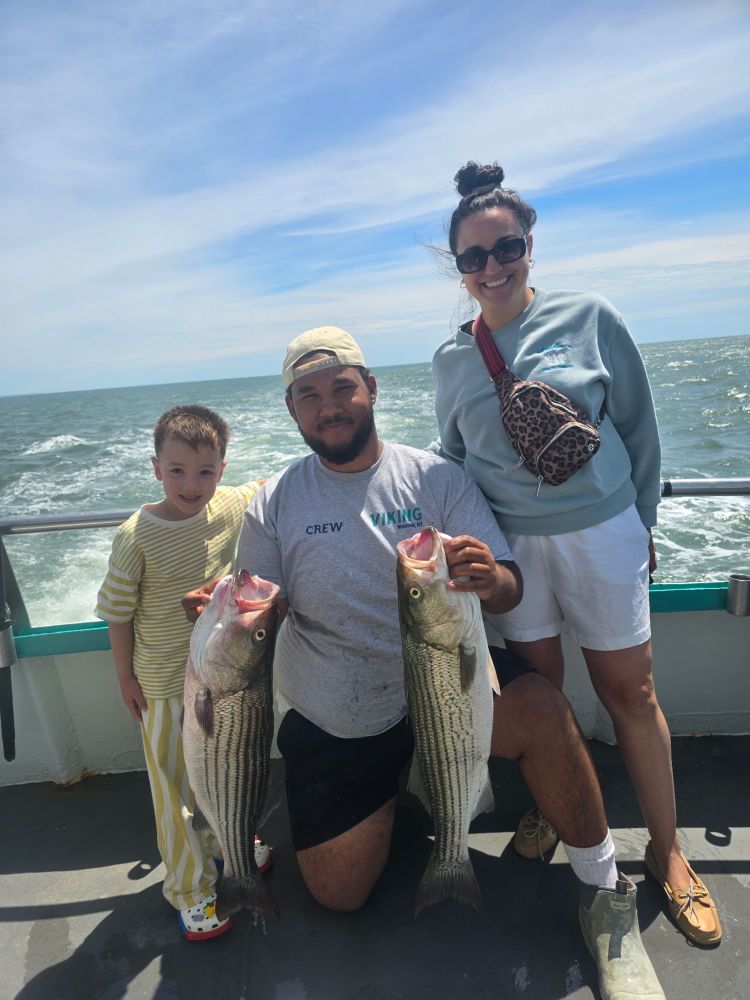 Two adults and a child on a boat, holding large fish, with ocean in the background.