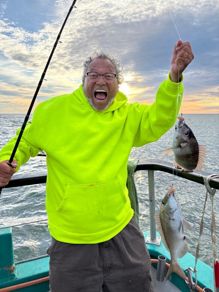 Person in bright hoodie holding two fish on a boat at sunset.