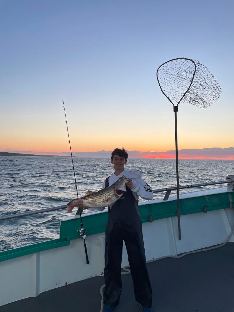 Person in overalls holding a large fish on a boat at sunset.