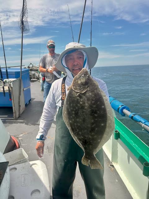 Person holding a flounder on a boat, another person in background.