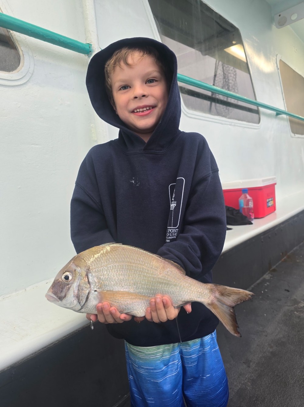Child in a hoodie holding a fish on a boat with a cooler in the background.