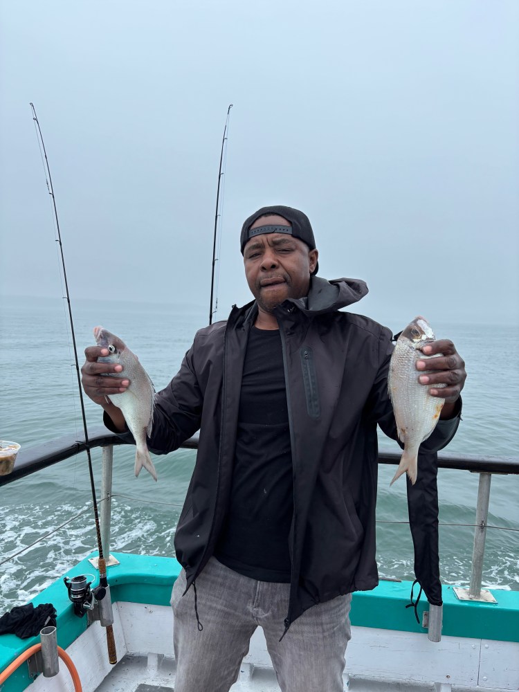 Person holding two fish on a boat, with fishing rods in the background.