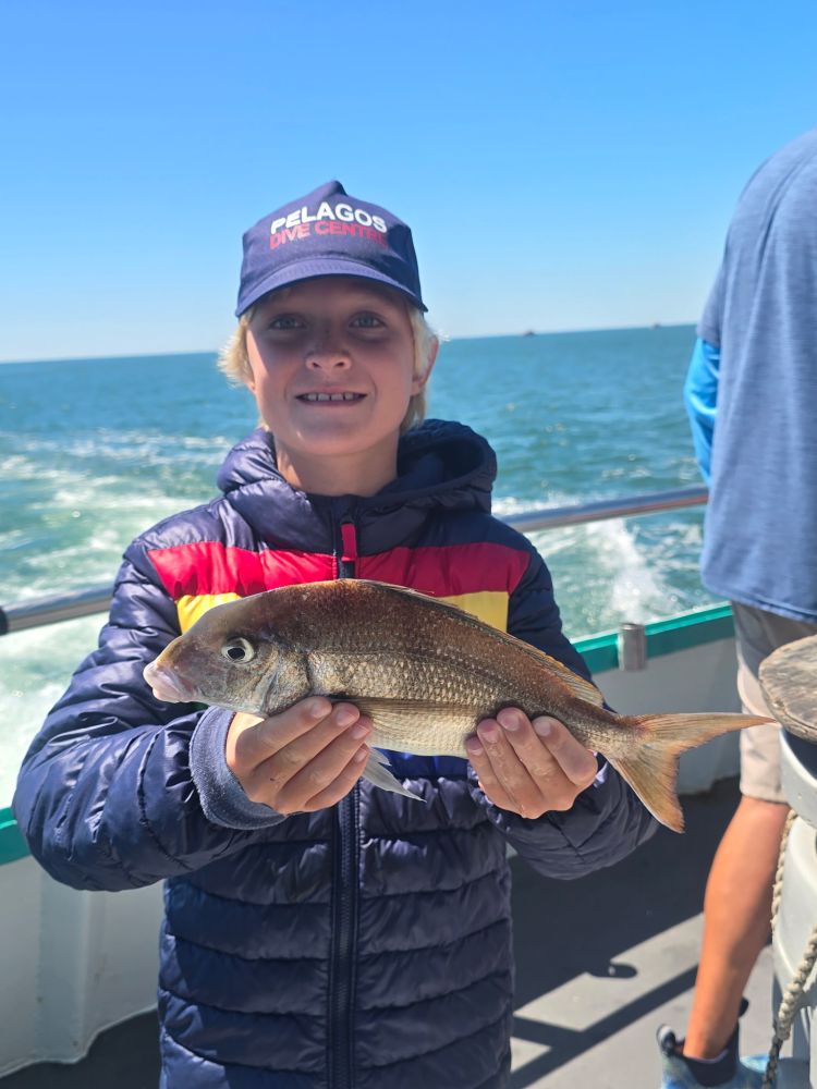 Child in a puffer jacket holds a fish on a boat with ocean in background.