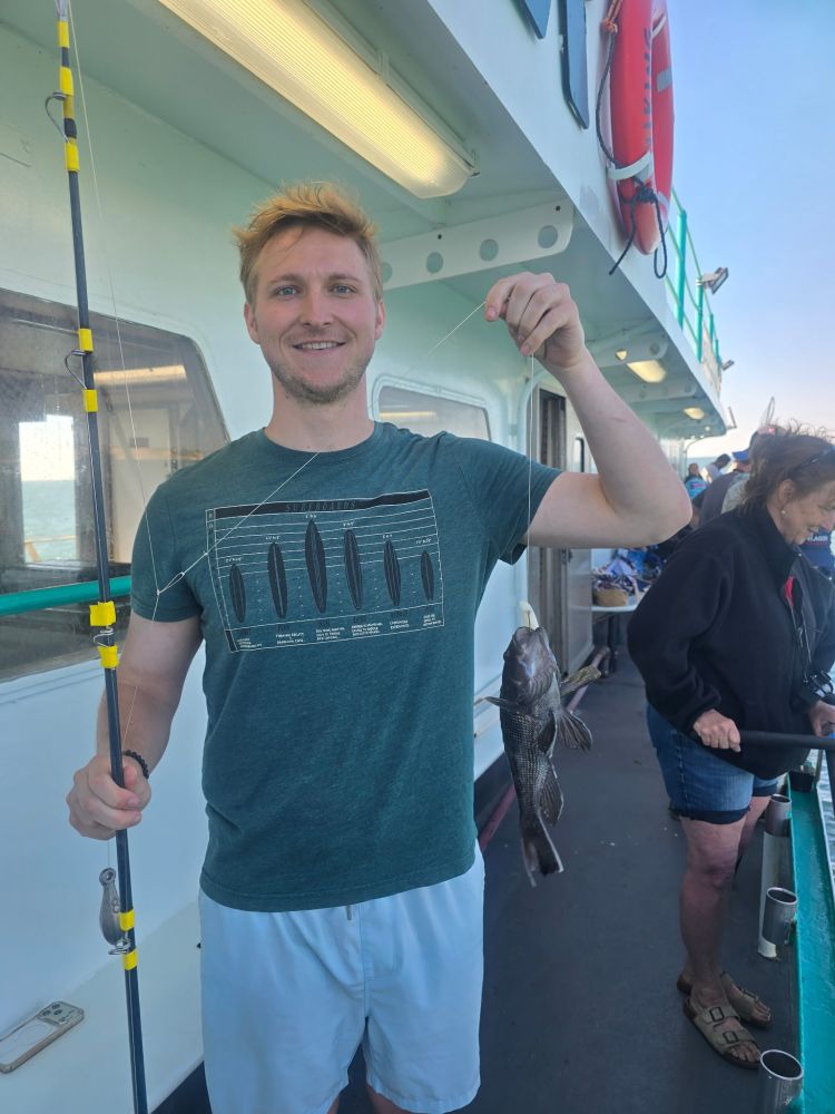 Person smiling, holding a small fish on a boat deck, with a fishing rod in the other hand.
