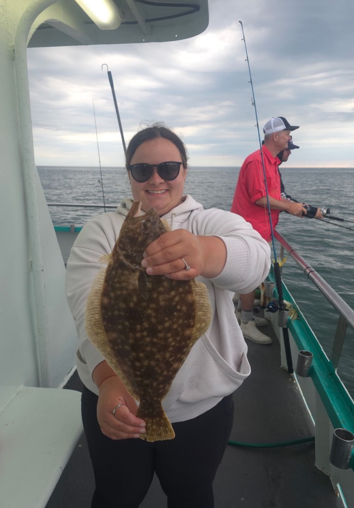 Person proudly holding a flatfish on a boat, with another person fishing in the background.